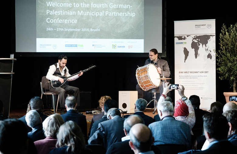 The photo shows two musicians on stage. The musician on the right in the picture holds a big drum. The musician on the left plays an oriental plucked instrument. In the background, a beamer has been projected onto the wall: 